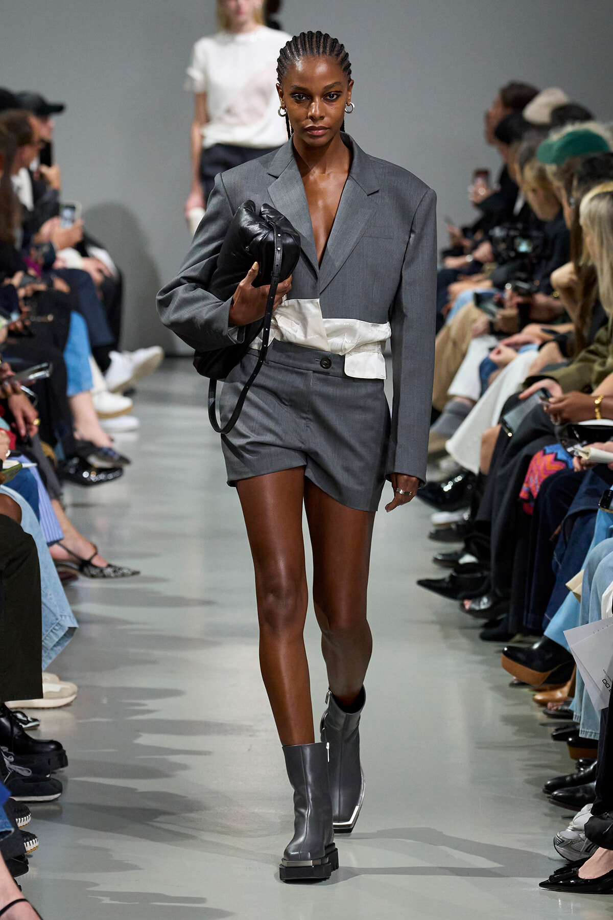 Runway model in a gray suit with blazer and matching shorts, carrying a black handbag as photographers and audience watch.