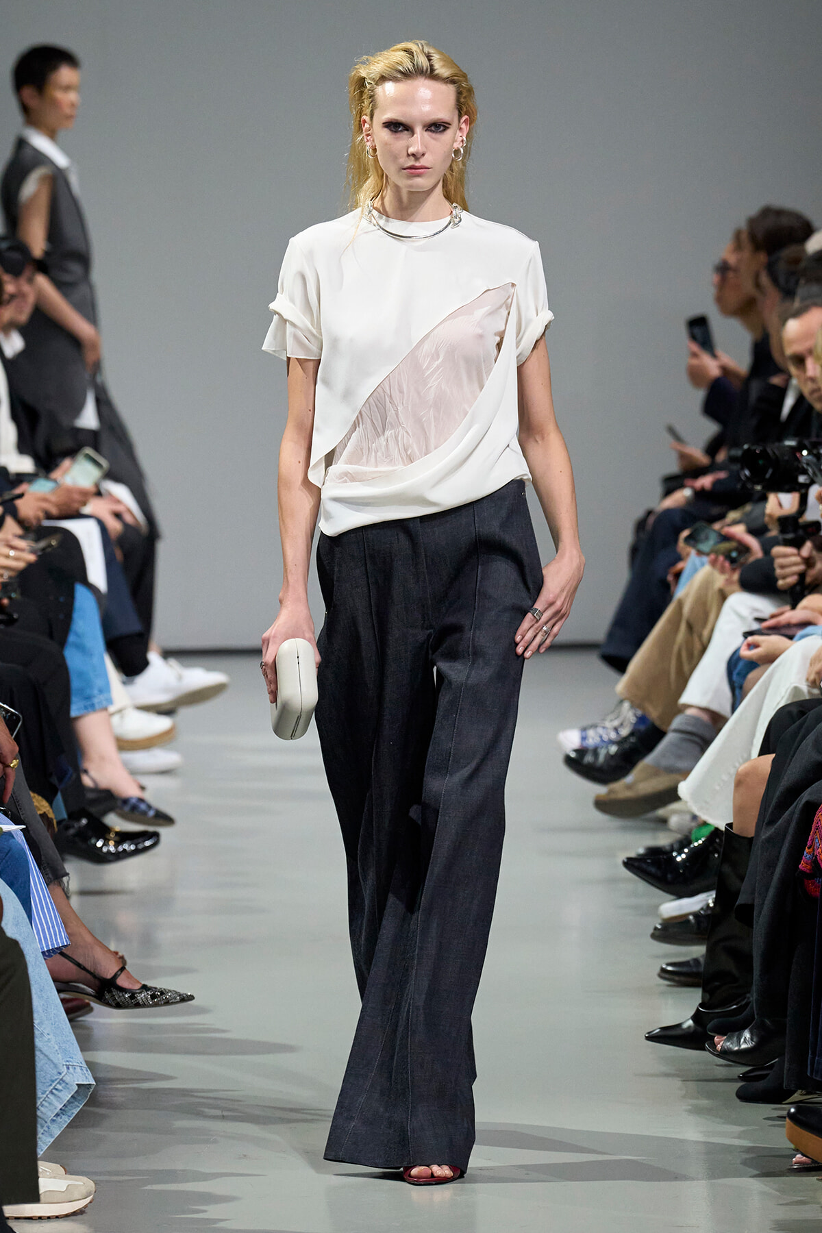 Model walks a fashion runway in a white draped top and wide-leg dark jeans, holding a small white clutch.