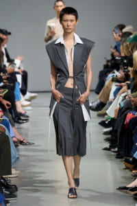 Model walks the runway in a gray sleeveless suit with a white wide-collar shirt, belt-tied at the waist.