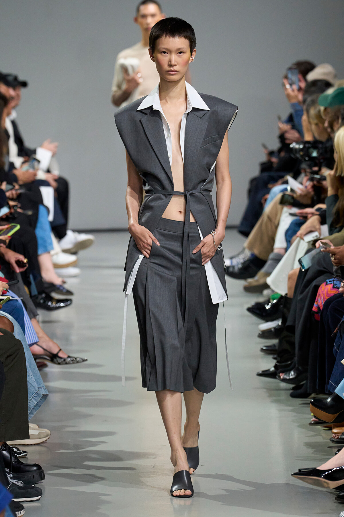 Model walks the runway in a gray sleeveless suit with a white wide-collar shirt, belt-tied at the waist.