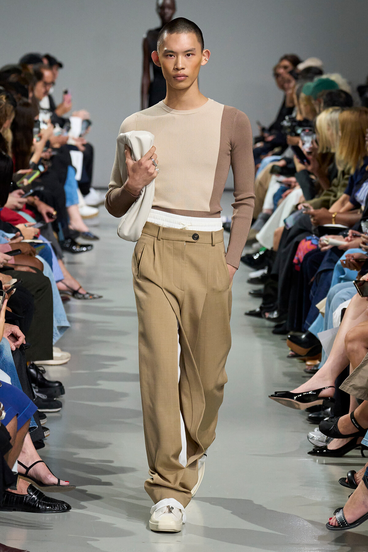 Male model walks the runway wearing a beige color-block knit top, tan pleated trousers, white sneakers, and carries a light bag.