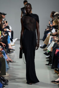 Model walks a black sheer top and full-length black skirt on a fashion runway, audience on both sides watching.