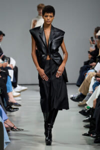 Model walks the runway in a black leather sleeveless blazer with a belt; spectators sit along the sides.Parallels: fashion show scene with a bold, all-black outfit.
