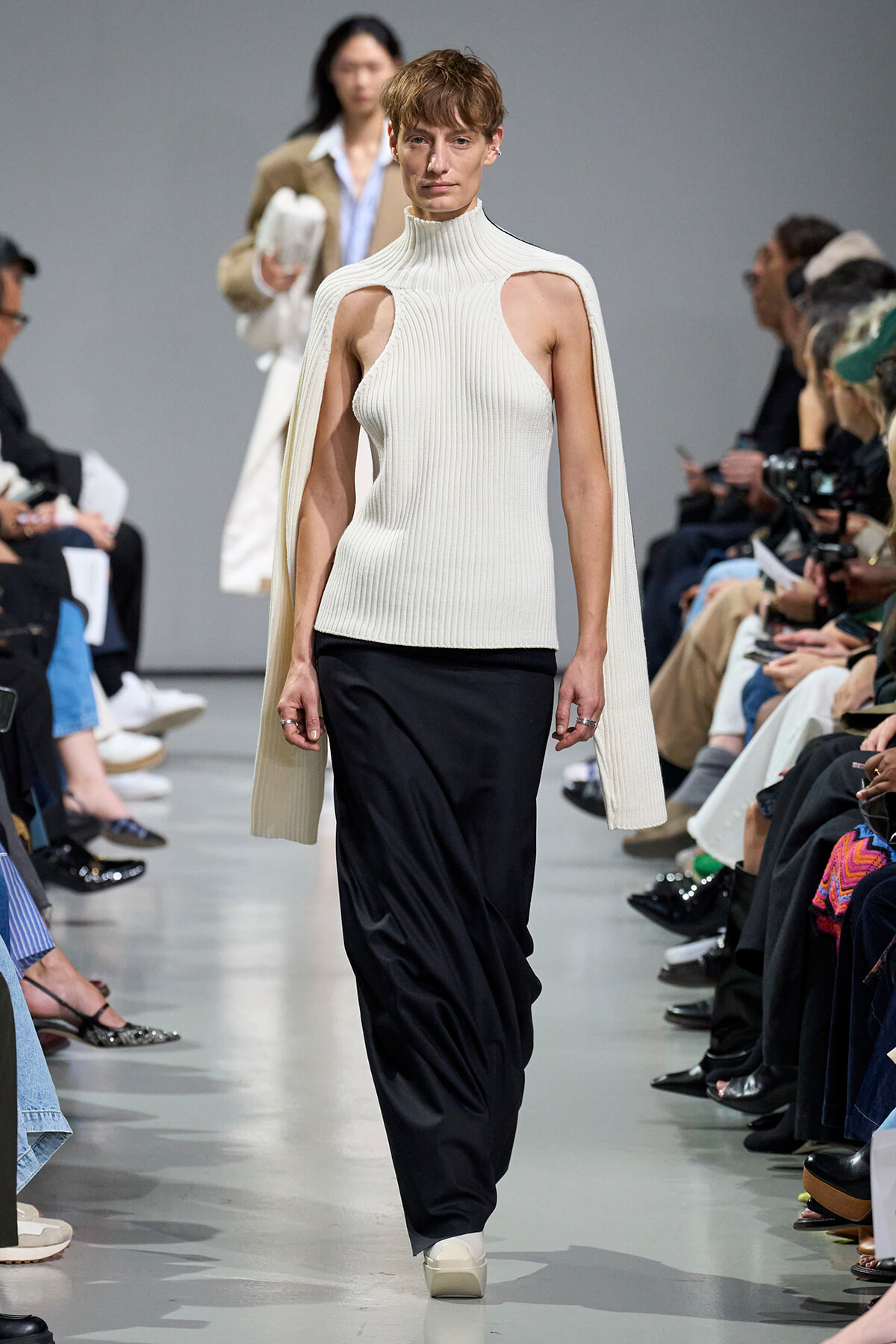 Model walks a runway in a cream ribbed sleeveless turtleneck with cape-like sleeves over black pants, audience on both sides.