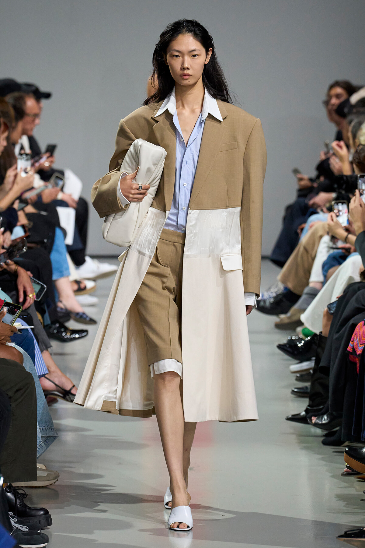 Model walks a fashion runway wearing a colorblock tan and cream oversized coat over a light blue shirt, carrying a white bag and white sandals amid seated spectators.