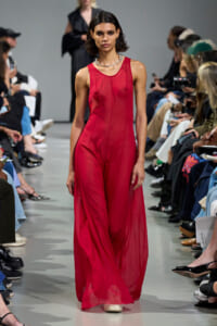 Model walks a fashion runway wearing a sleeveless red sheer jumpsuit and a chunky silver necklace, audience on both sides.