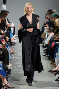 Model walks the runway in a long, black, layered trench coat with oversized sleeves.","Female model wearing an oversized black coat with dramatic, wide lapels on a catwalk.","Runway moment: a model in a voluminous black trench strides toward the camera as the audience watches.","Fashion show image of a model in a black, floor-length coat with strong structure.","Model crosses the catwalk in a dark, dramatic black coat; audience seated on both sides."]} }*** wait there is a syntax error: I added stray quotes and not proper JSON. Ensure valid JSON. Also the content should be enclosed correctly. Let's fix: The array items should be strings without stray quotes inside. Also one extra quote after period. Let's craft proper. We'll deliver correct JSON. We'll ensure no trailing stray. Also maybe 4 options is fine. Provide 5. Let's compose. } Let's produce. } Let's finalize. Ensure altTexts array. } Let's output. }? Provide only the JSON as required. ``` JSON. Remove code blocks. The required format: {