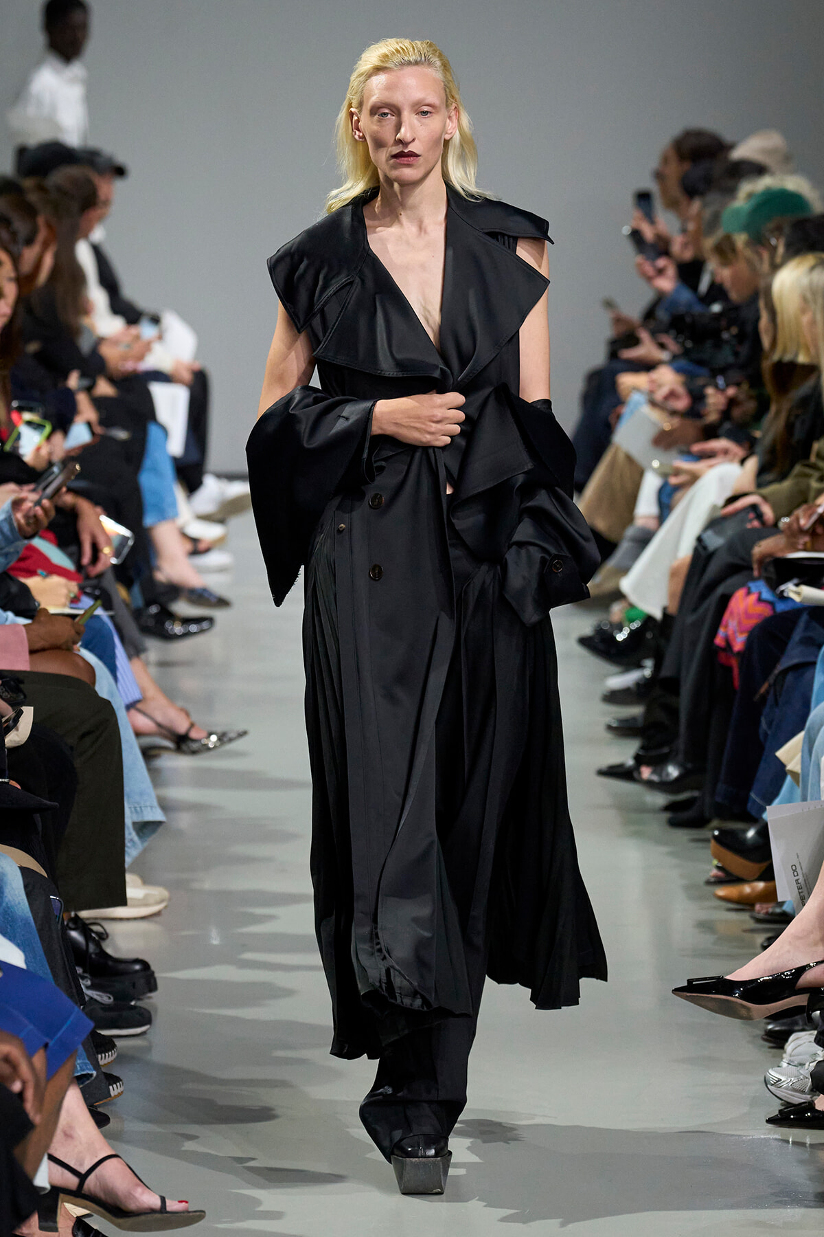 Model walks the runway in a long, black, layered trench coat with oversized sleeves.","Female model wearing an oversized black coat with dramatic, wide lapels on a catwalk.","Runway moment: a model in a voluminous black trench strides toward the camera as the audience watches.","Fashion show image of a model in a black, floor-length coat with strong structure.","Model crosses the catwalk in a dark, dramatic black coat; audience seated on both sides."]} }*** wait there is a syntax error: I added stray quotes and not proper JSON. Ensure valid JSON. Also the content should be enclosed correctly. Let's fix: The array items should be strings without stray quotes inside. Also one extra quote after period. Let's craft proper. We'll deliver correct JSON. We'll ensure no trailing stray. Also maybe 4 options is fine. Provide 5. Let's compose. } Let's produce. } Let's finalize. Ensure altTexts array. } Let's output. }? Provide only the JSON as required. ``` JSON. Remove code blocks. The required format: {