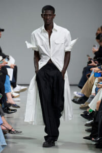 Male model walking a fashion runway in a white shirt and long white coat over black pants, audience on both sides watching