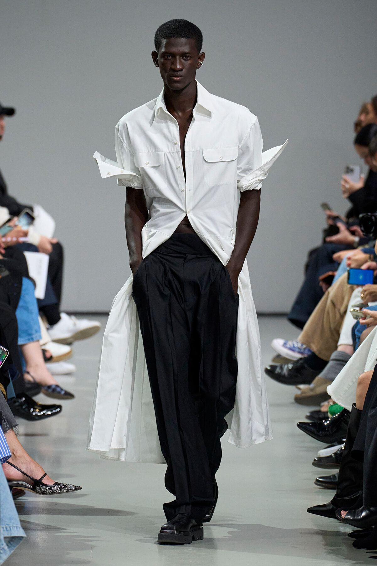 Male model walking a fashion runway in a white shirt and long white coat over black pants, audience on both sides watching