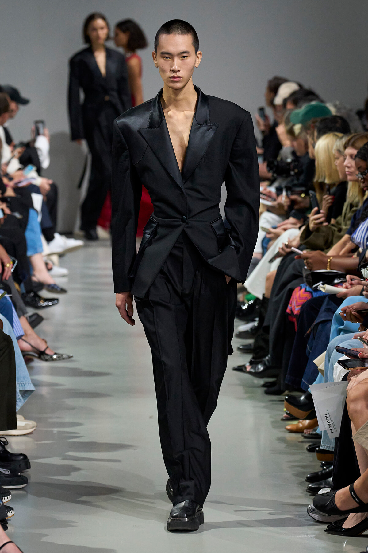 Male model walks the runway in a black satin suit with an open deep V-neck and wide lapels; audience seated on both sides watches the show.