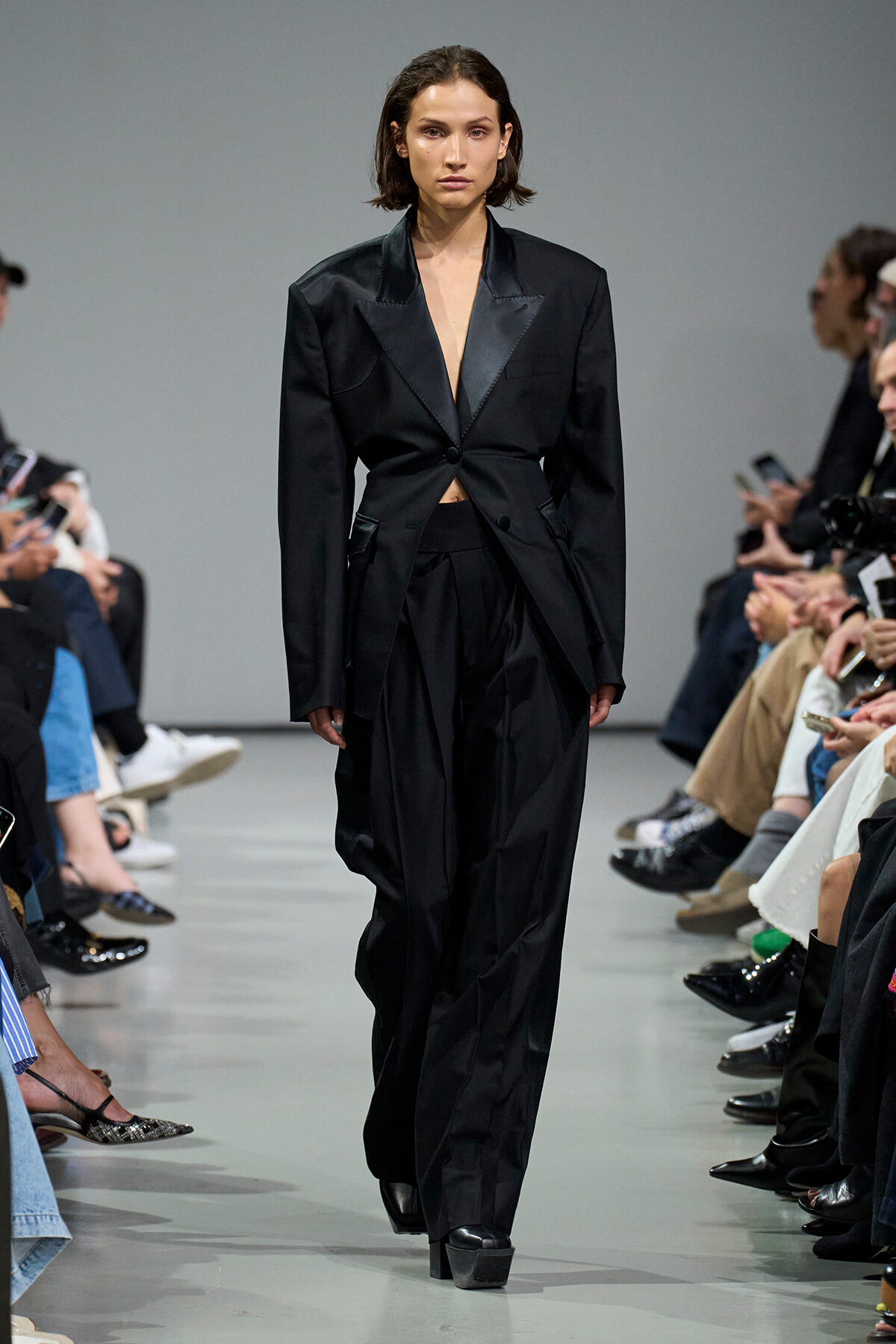 Model walks a fashion runway wearing a black, tailored suit with a deep V-neck lapel, audience seated along the sides.