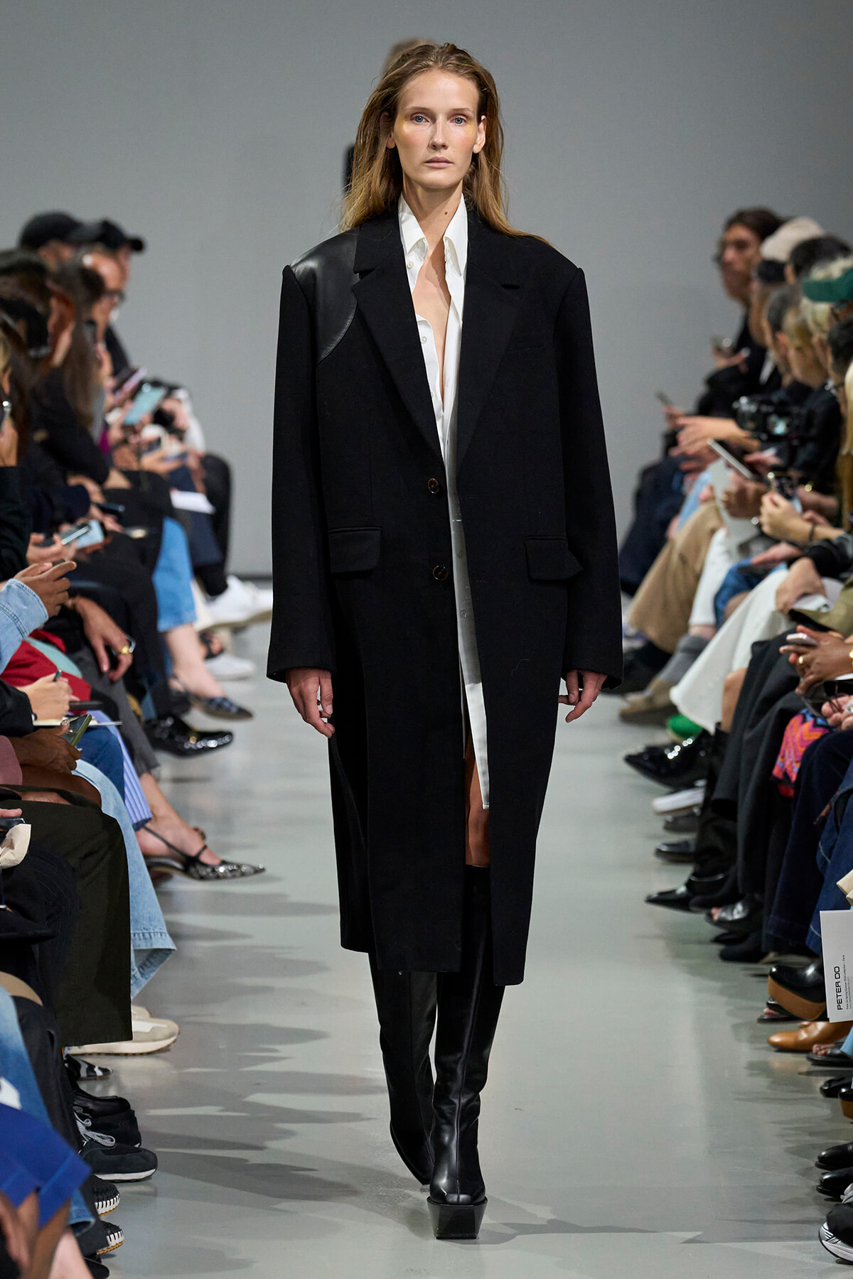 Female model walks a fashion runway in a long black coat over a white shirt, with spectators on both sides.