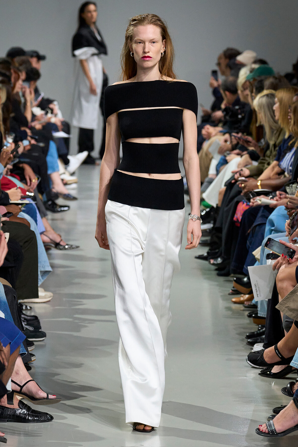 Model walking a fashion runway in a black off-the-shoulder crop top and white wide-leg pants, audience on both sides watching
