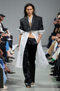 Model walks the runway in an oversized black blazer layered over a white shirt with a long white shirt coat and black pants.