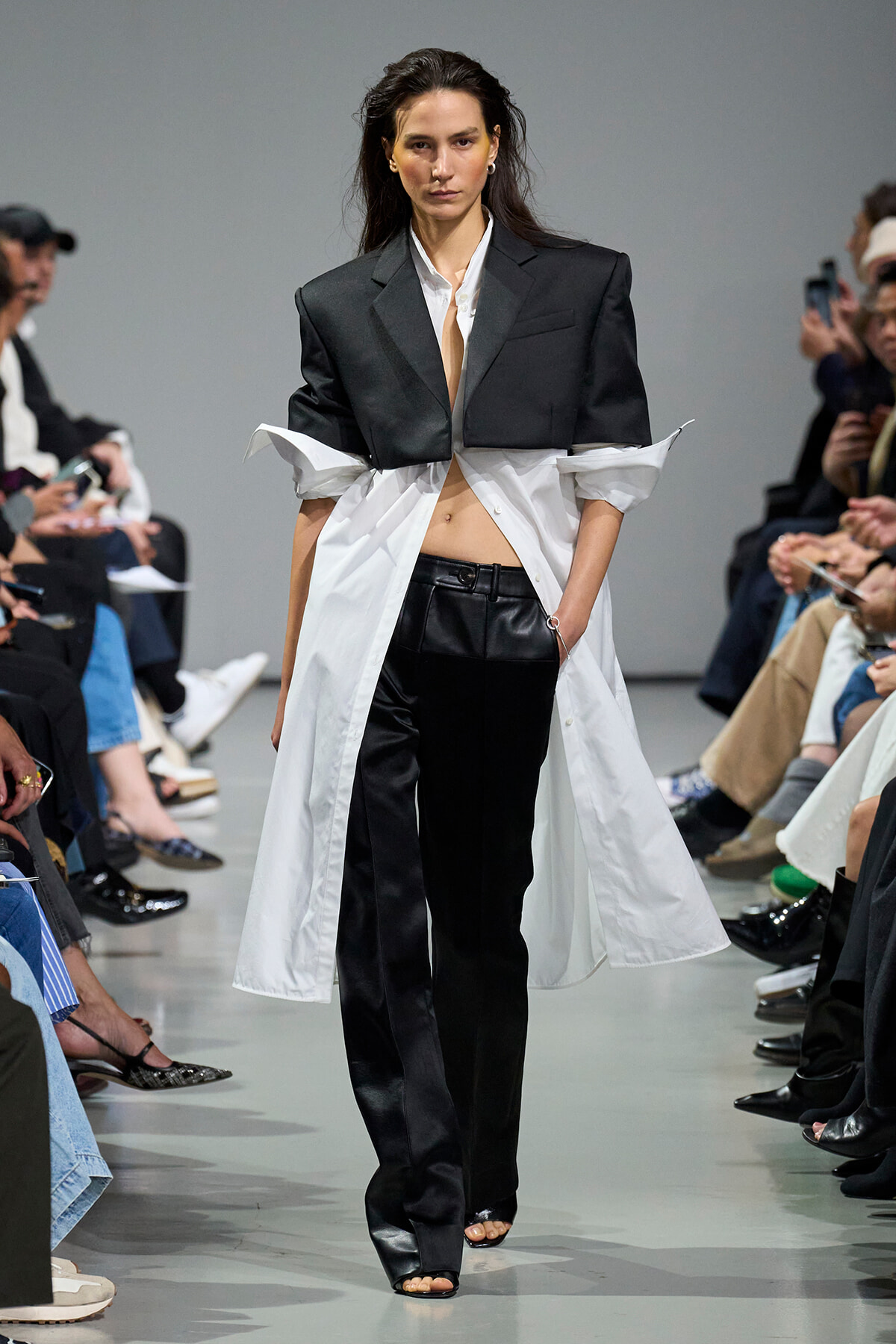Model walks the runway in an oversized black blazer layered over a white shirt with a long white shirt coat and black pants.