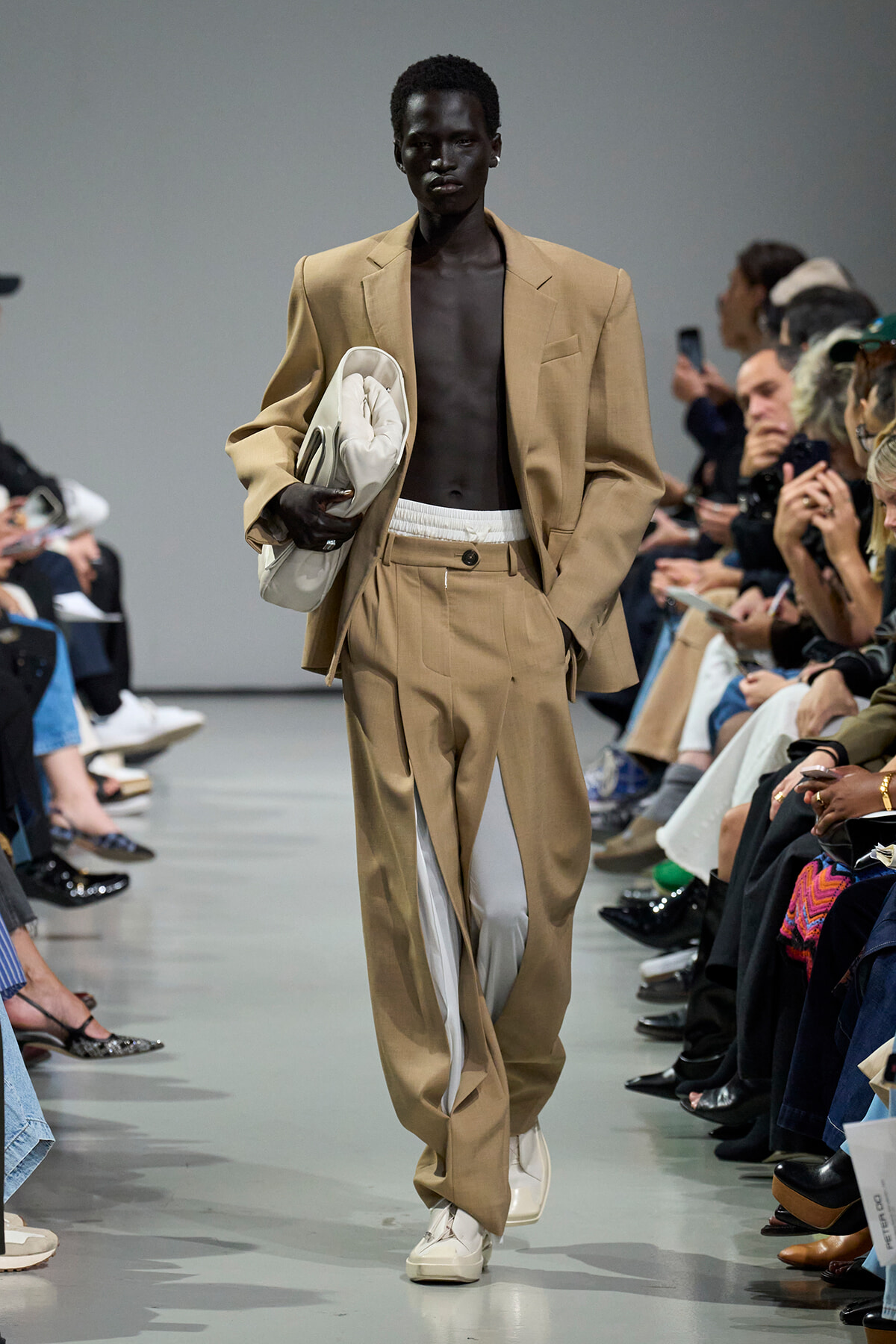 Male model walks the runway in a tan suit wearing white sneakers, carrying a white garment bag, with audience on both sides.