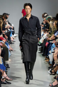 Model in an all-black outfit—a button-down shirt, high-waisted pants, and knee-high boots—walks the runway with seated audience on both sides.