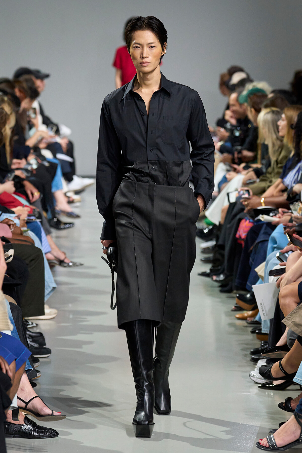 Model in an all-black outfit—a button-down shirt, high-waisted pants, and knee-high boots—walks the runway with seated audience on both sides.