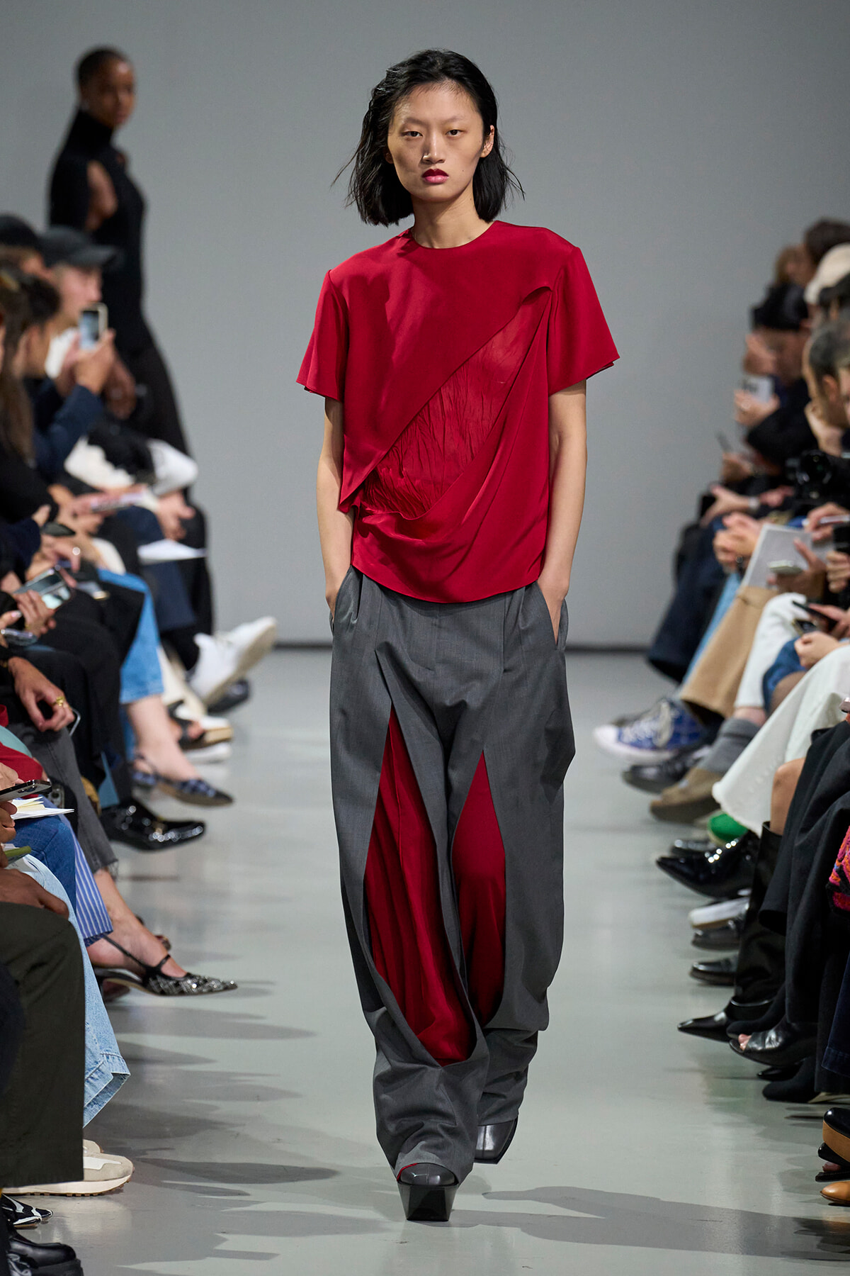 Model walks the runway in a red draped top paired with gray, wide-leg pants showing red lining, audience on either side.