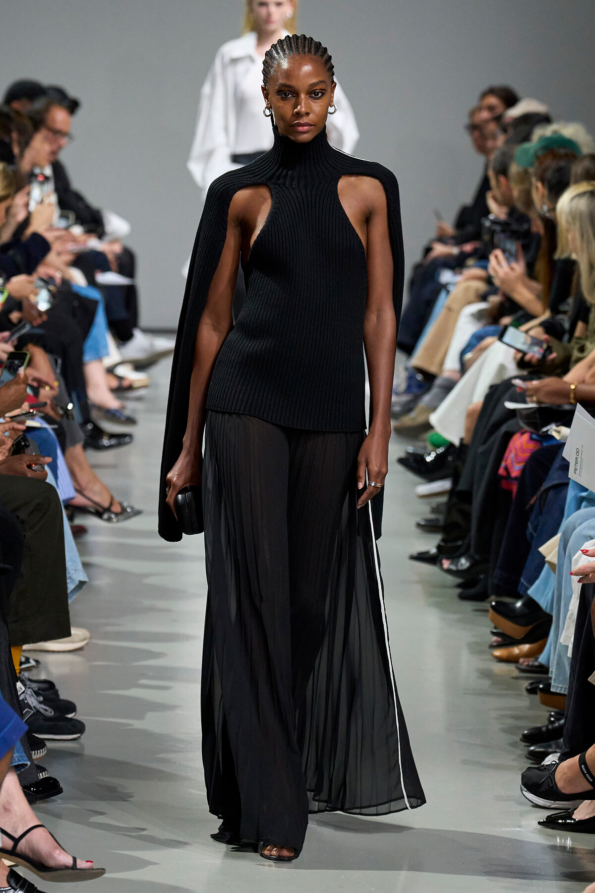 Female model walks a runway in a black ribbed knit dress with cutouts at the shoulders, paired with a sheer, floor-length skirt and cape-like sleeves, audience on both sides photographing the scene.