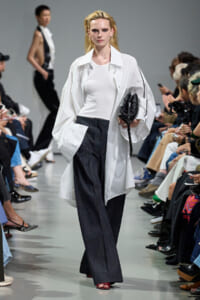 Model on a fashion runway in a white oversized jacket over a white top and dark wide-leg pants, carrying a black clutch.