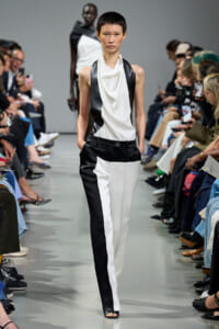 Model walking the runway in a black-and-white sleeveless draped top and color-block pants; audience lines the catwalk behind.