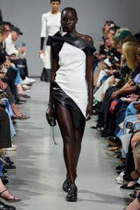 Model walks a runway in a black-and-white asymmetric dress as photographers and spectators line the catwalk.
