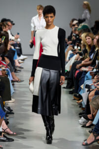 Model walking a fashion runway in a color-block knit top and black leather skirt, audience on both sides watching.
