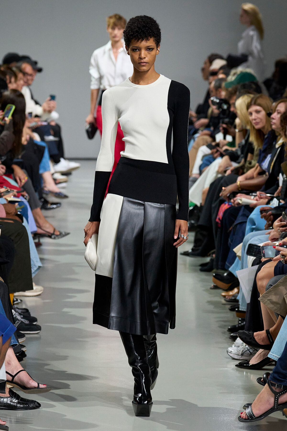 Model walking a fashion runway in a color-block knit top and black leather skirt, audience on both sides watching.