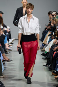Model walks the runway in a white button-down shirt and sheer red pants with a black high-waisted band; audience flanks the catwalk.