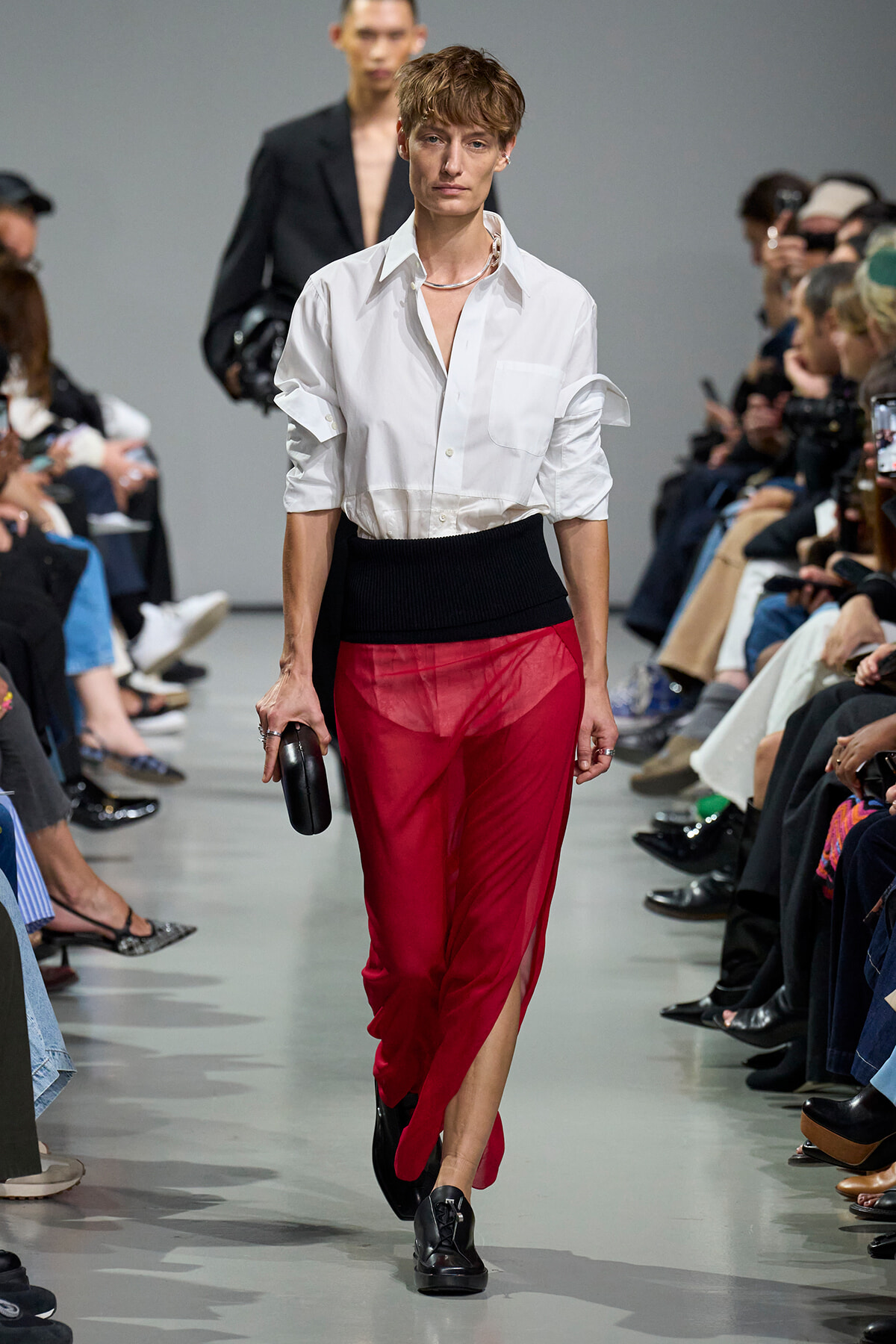 Model walks the runway in a white button-down shirt and sheer red pants with a black high-waisted band; audience flanks the catwalk.