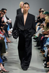 Model walks the runway in a black oversized suit with a deep neckline, carrying a black clutch as spectators watch from both sides.