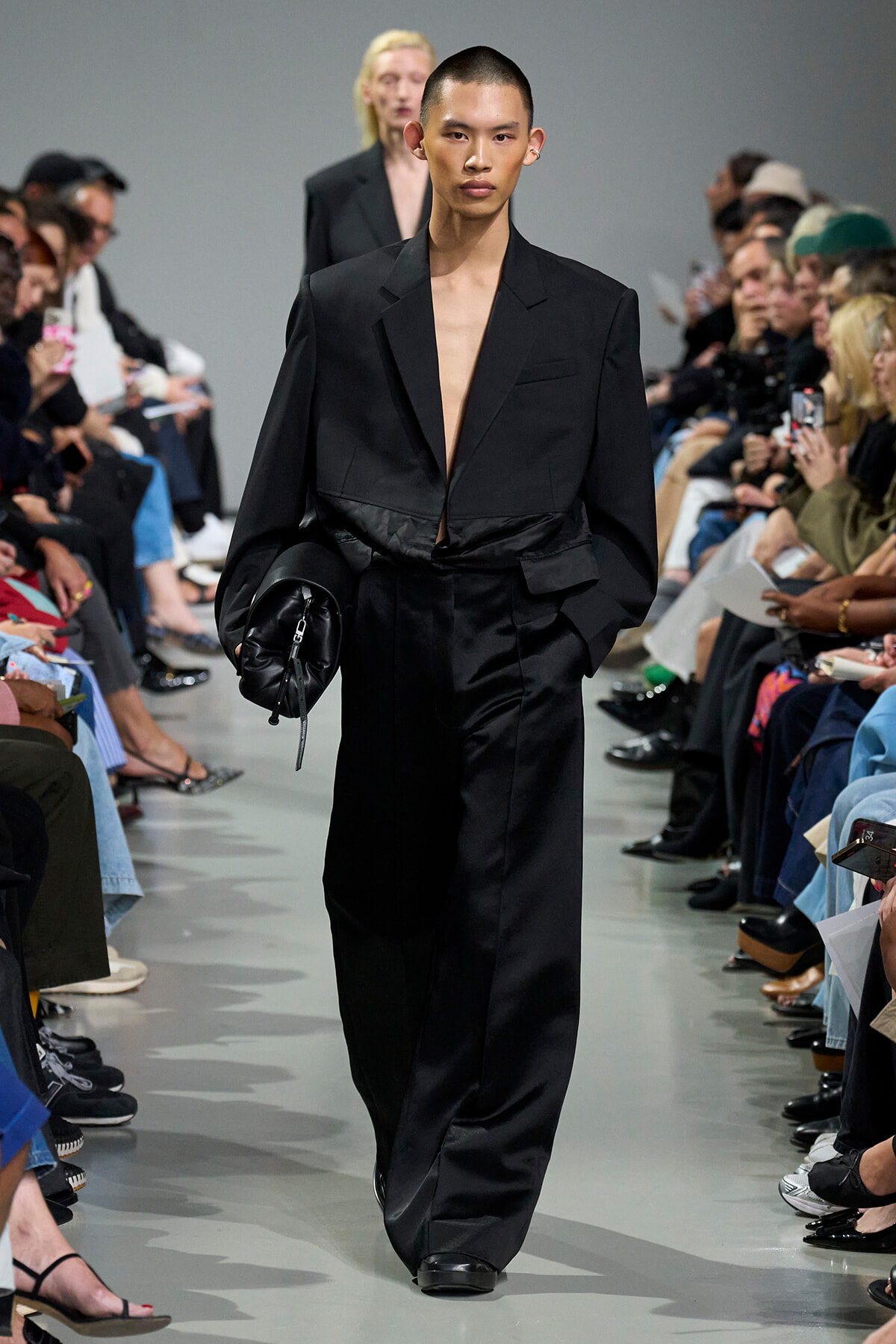 Model walks the runway in a black oversized suit with a deep neckline, carrying a black clutch as spectators watch from both sides.