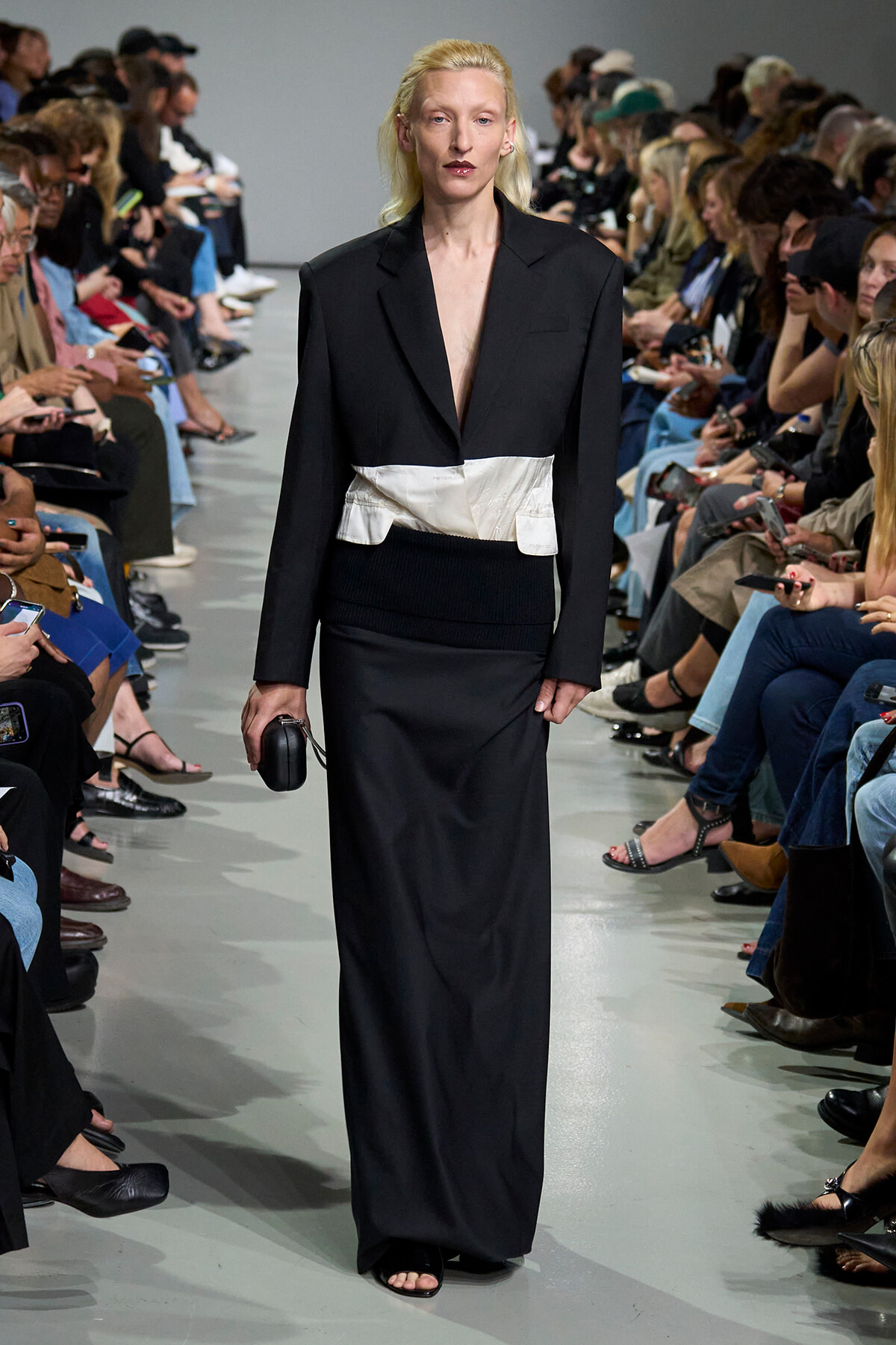 Model walks the runway in a black tailored blazer dress with a white waist wrap, audience seated on both sides watching.
