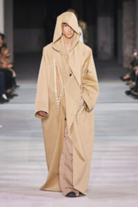 Male model walking a runway in a long beige trench with oversized, draped hooded design.