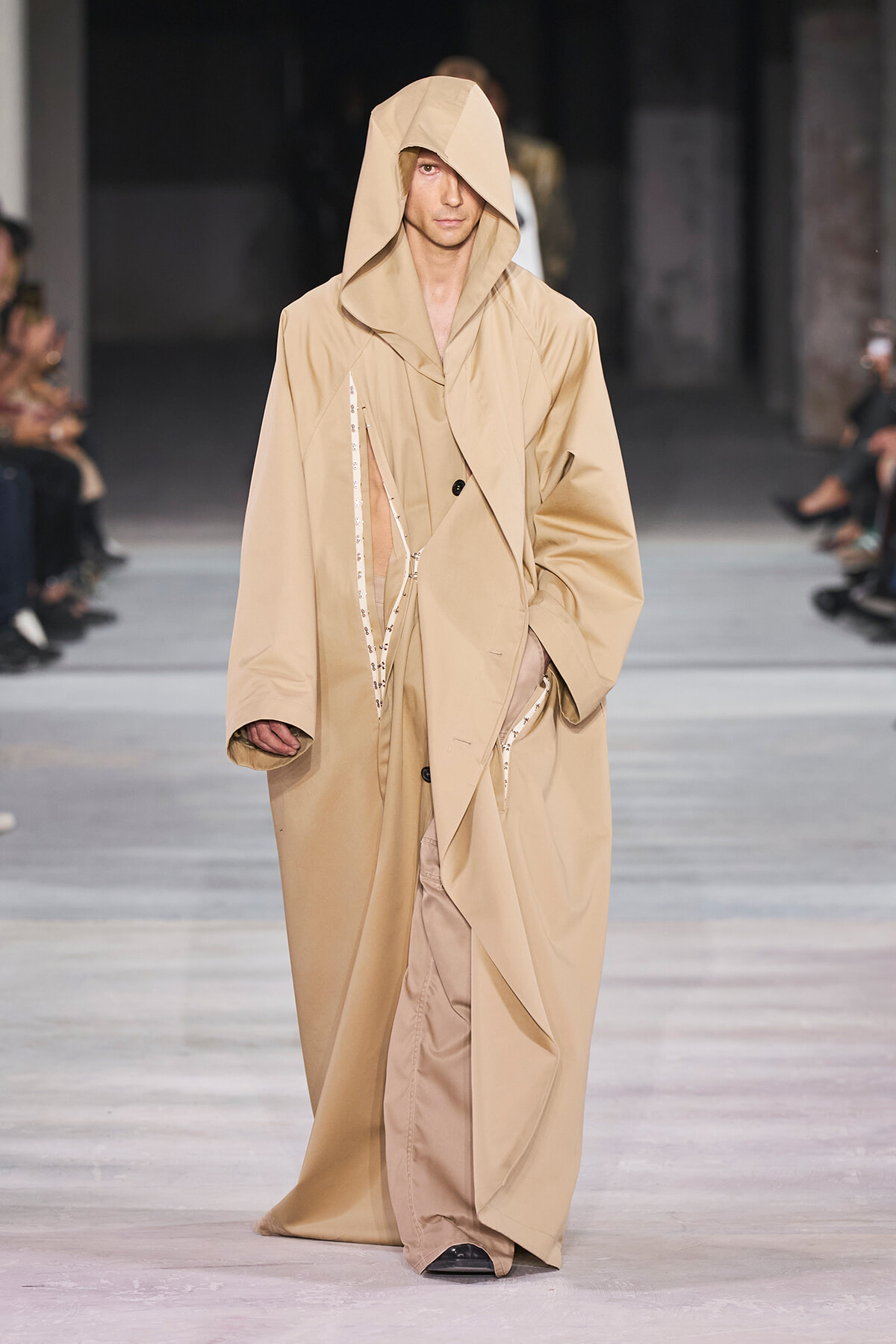 Male model walking a runway in a long beige trench with oversized, draped hooded design.