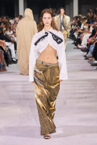 Model on a runway wearing a white twisted-top blouse and shimmering gold wide-leg pants with spectators along the catwalk.