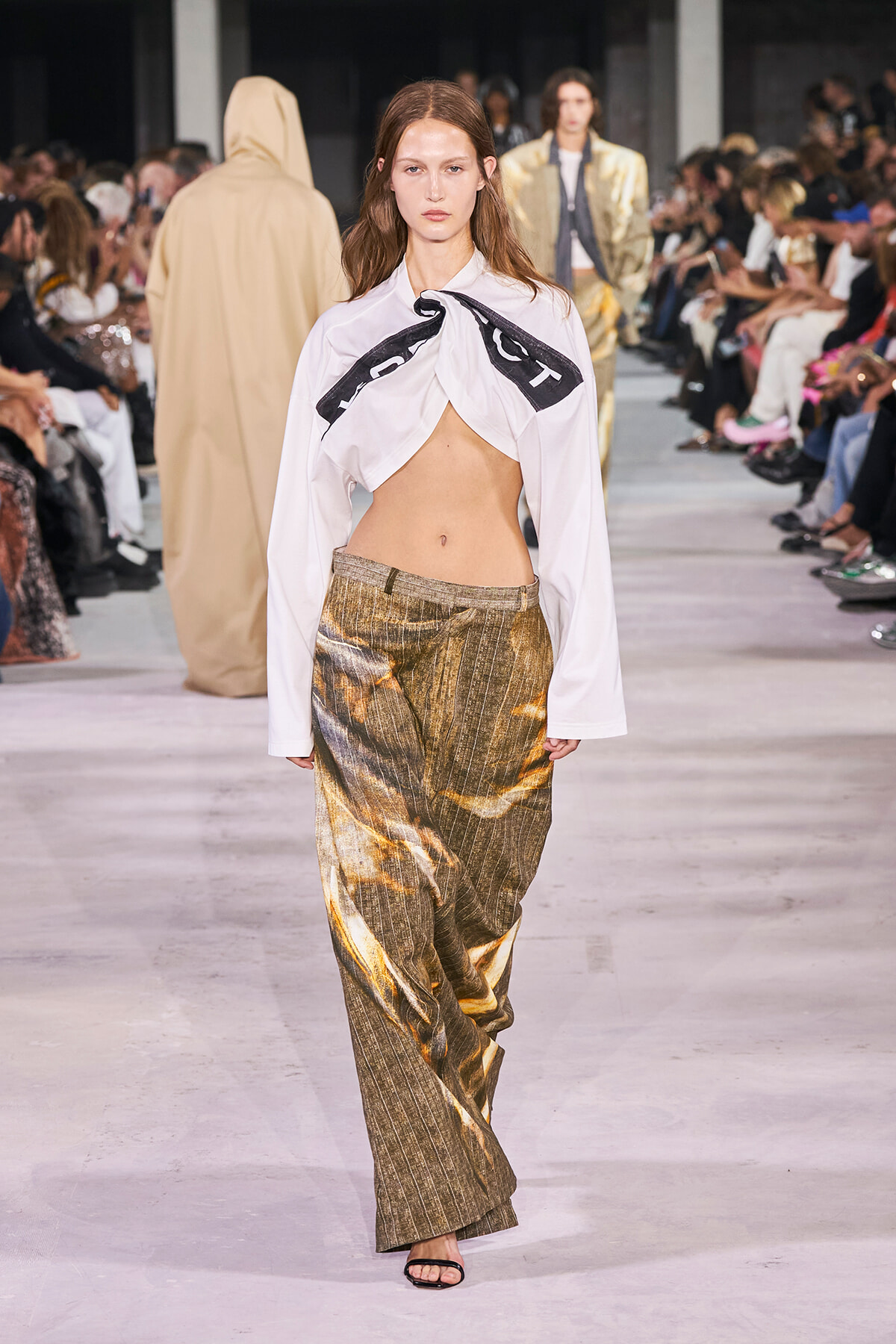 Model on a runway wearing a white twisted-top blouse and shimmering gold wide-leg pants with spectators along the catwalk.