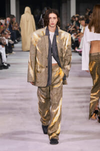 Male model in a gold metallic pinstripe suit with a gray inner layer walks the runway; audience visible on both sides.