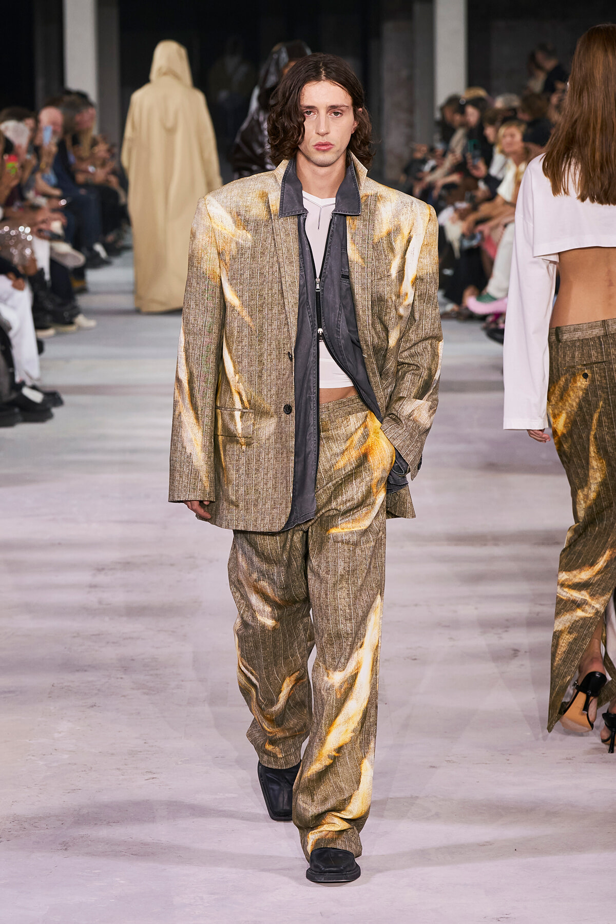 Male model in a gold metallic pinstripe suit with a gray inner layer walks the runway; audience visible on both sides.