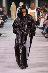 Model walks the runway in a long, shiny black hooded coat with an oversized fit.