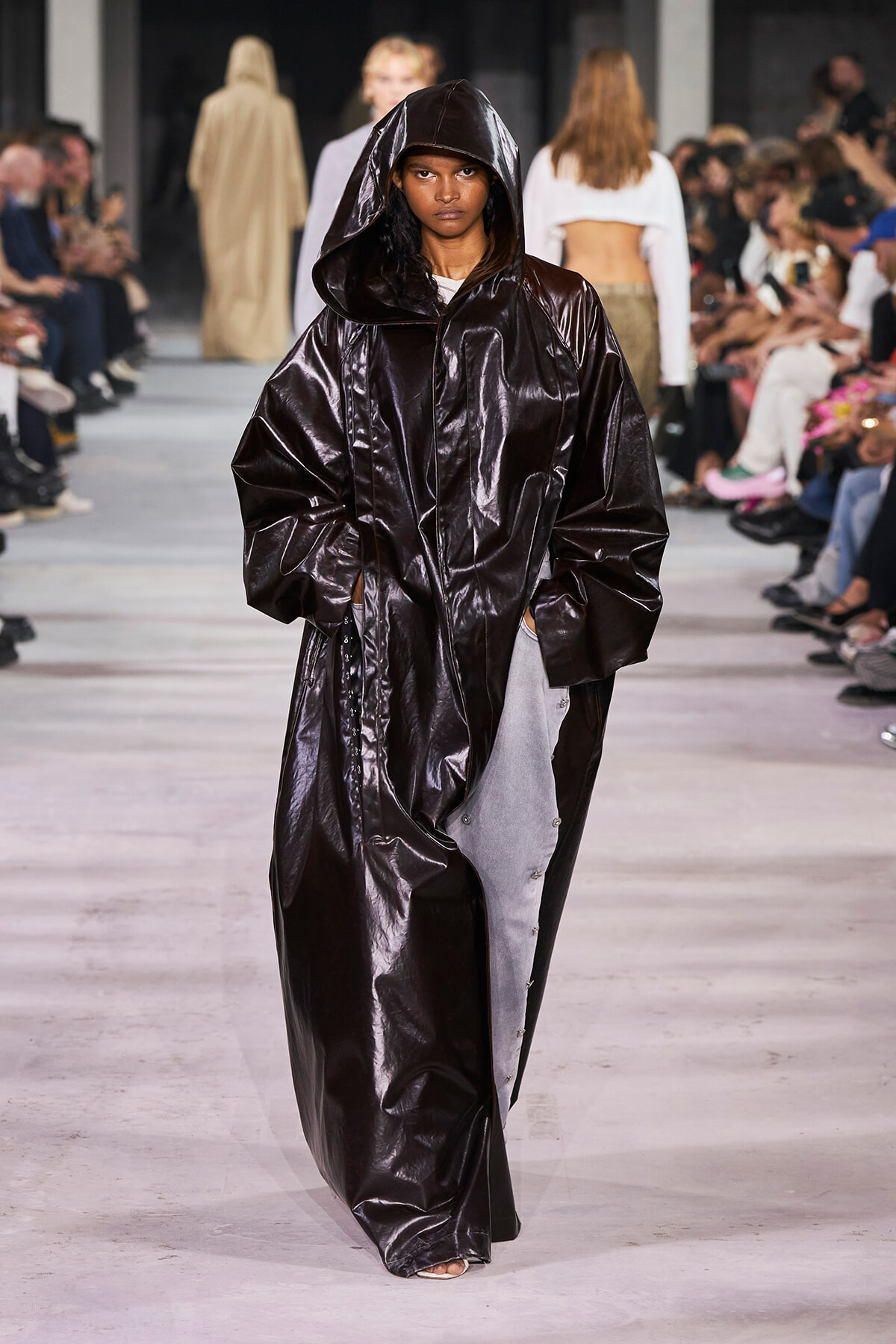 Model walks the runway in a long, shiny black hooded coat with an oversized fit.