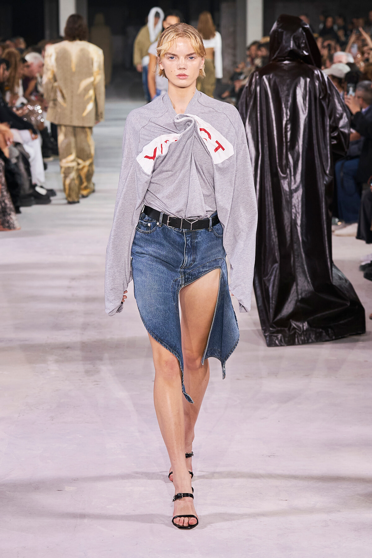 Model walks a fashion runway in a light gray twisted-top blouse tucked into a high-slit denim skirt with a black belt and strappy sandals; audience blurred in background.
