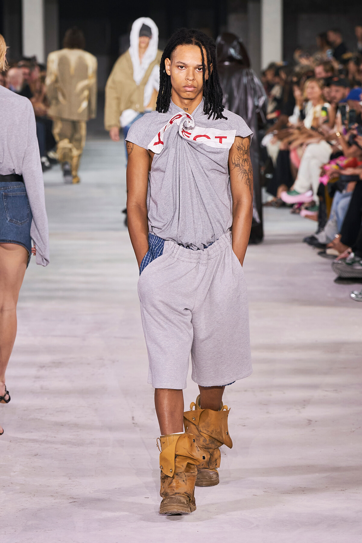 Model walks a fashion runway in a light gray, knotted tee and matching shorts; tan boots and a crowd of spectators line the catwalk.