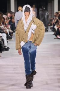 Model walks a fashion show in an oversized tan bomber jacket over a light gray hoodie, cap, and blue jeans with button details, black boots.