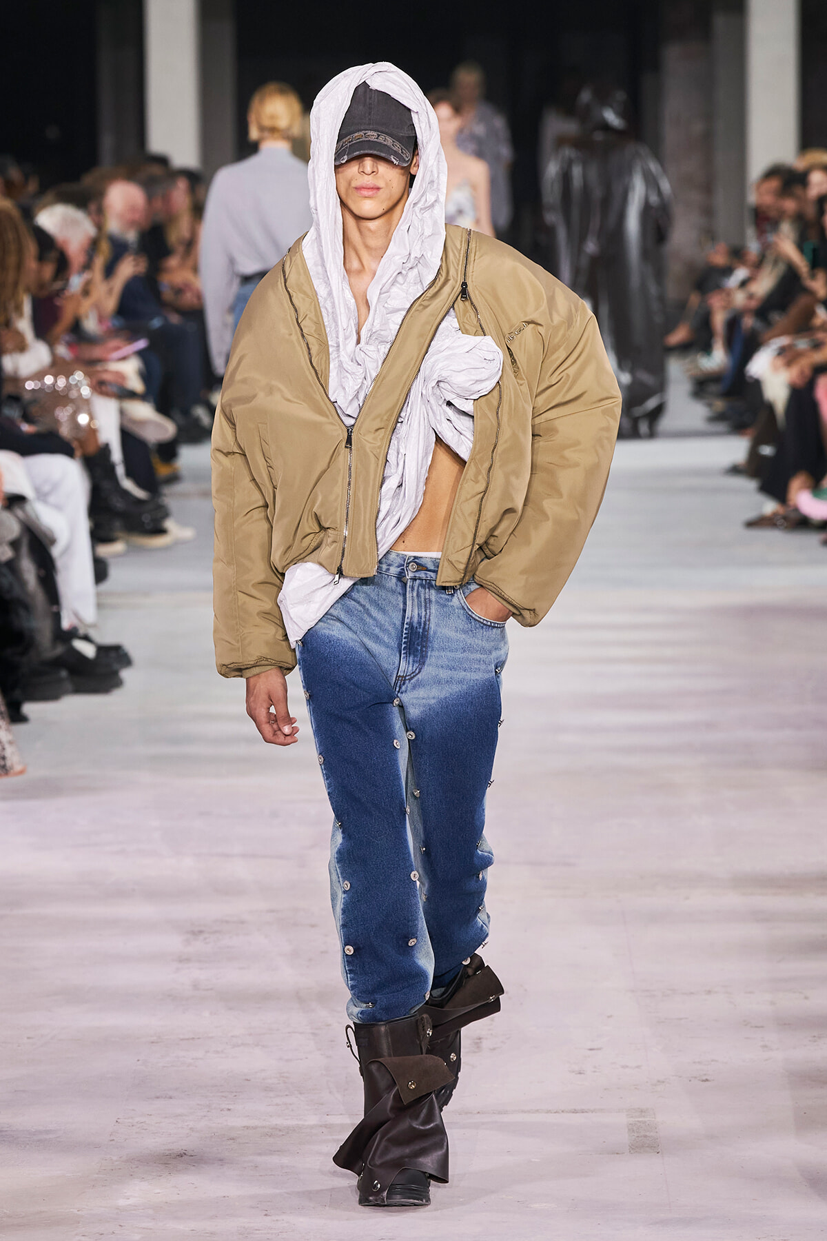 Model walks a fashion show in an oversized tan bomber jacket over a light gray hoodie, cap, and blue jeans with button details, black boots.