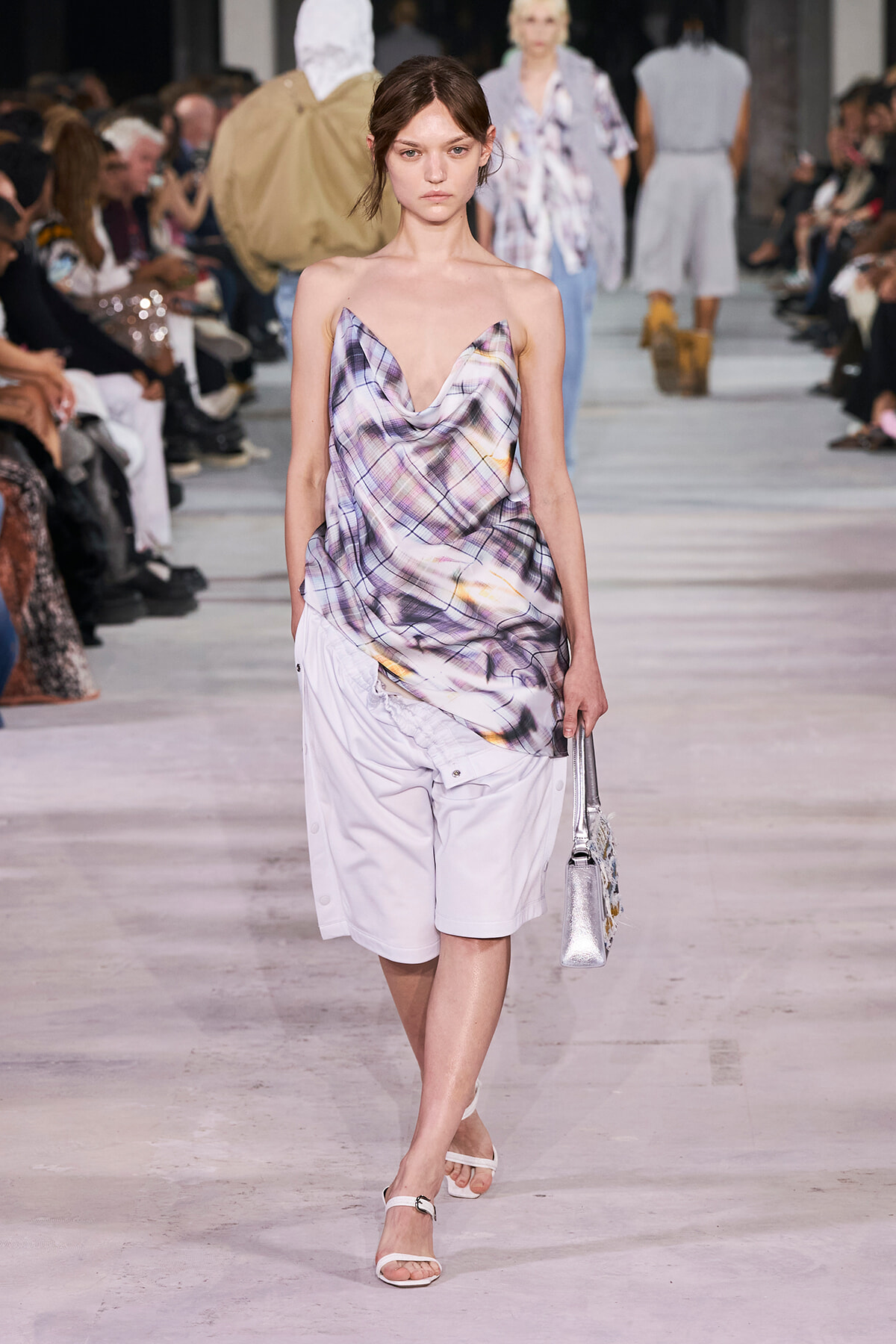Model walks the runway in a draped lavender plaid top and white shorts, carrying a silver handbag.