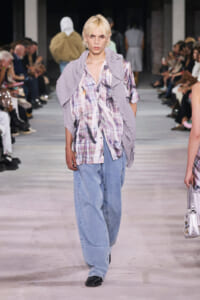 Fashion model walking a runway in a lavender cardigan draped over a pastel plaid shirt and wide light-blue jeans, with audience on both sides.