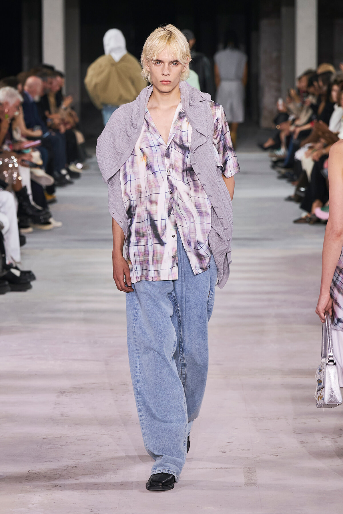 Fashion model walking a runway in a lavender cardigan draped over a pastel plaid shirt and wide light-blue jeans, with audience on both sides.